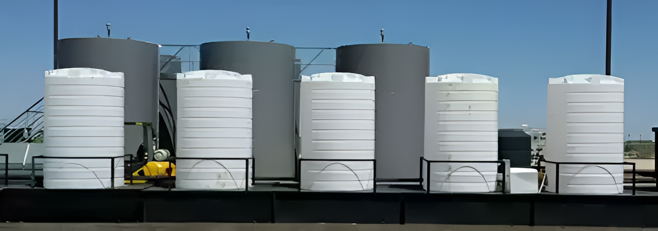 Multiple white vertical poly tanks lined up in front of larger industrial storage tanks